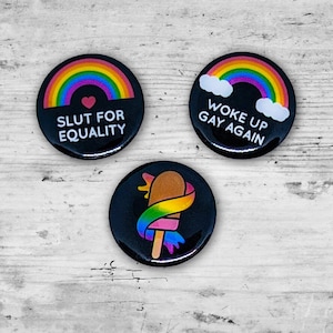 May include: Three black round buttons with rainbow designs. One button says "SLUT FOR EQUALITY" with a rainbow and heart. Another says "WOKE UP GAY AGAIN" with a rainbow and clouds. The third button features a popsicle wrapped in a rainbow ribbon.