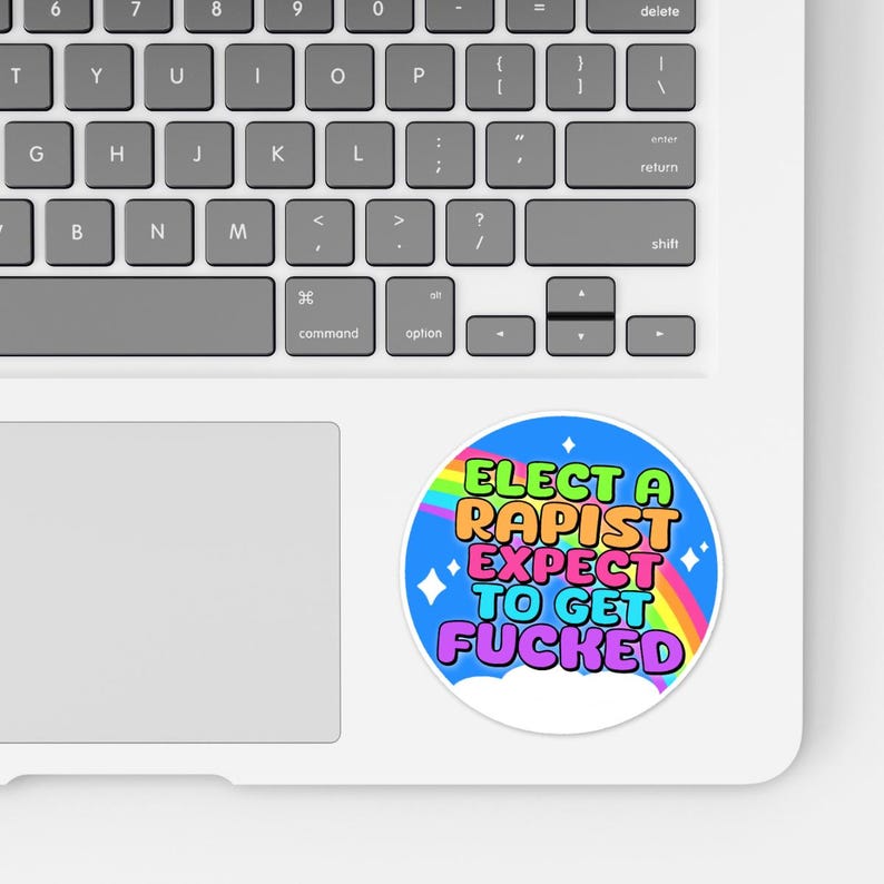 May include: A round sticker with the text "ELECT A RAPIST EXPECT TO GET FUCKED" in bright colors. The sticker has a blue background with a rainbow and stars. The sticker is on a white laptop.