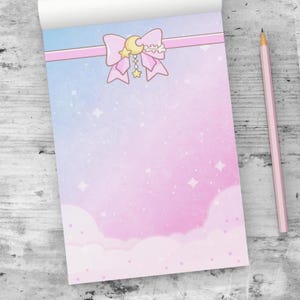 May include: A notepad with a pastel pink and blue watercolor design. The top features a pink ribbon and a bow with a crescent moon and stars. A pink pencil rests on the side. The notepad has a cloud and star pattern.