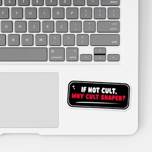 May include: A white laptop with a sticker that reads "IF NOT CULT, WHY CULT SHAPED?" in red and white text on a black background. The sticker is rectangular with rounded corners. The laptop keyboard is visible.
