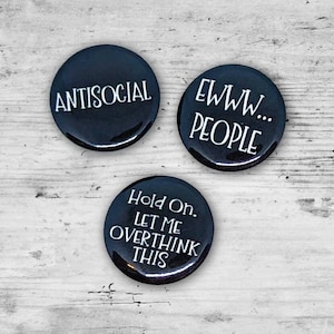 May include: Three black round buttons with white text. One button says "ANTISOCIAL", another says "EWWW... PEOPLE", and the third says "Hold On. LET ME OVERTHINK THIS". The buttons are on a white wooden surface.