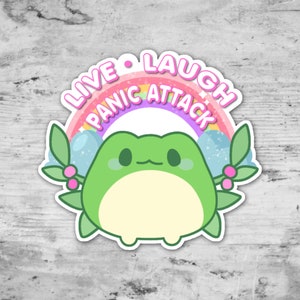 May include: A green frog with a pink rainbow above it that says "Live Laugh Panic Attack". The frog is surrounded by green leaves with pink berries.