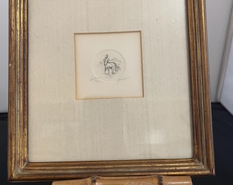 May include: A framed artwork featuring a small circular image of a person on a horse, with script below. The frame is gold-colored with a textured surface. The artwork is displayed on a bamboo easel.
