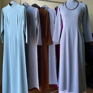 May include: A row of long-sleeved dresses in various colors, including light blue, gray, and brown, hanging on a clothing rack. Each dress has a mandarin collar and a long, flowing silhouette. One dress has a necklace.
