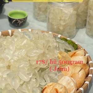 May include: A close-up of a clear glass jar filled with dried palm fruit pieces. The jar is sitting on a woven basket with other pieces of dried palm fruit. The text "17$/ hũ 500gram (Jolm)" is visible in the image.