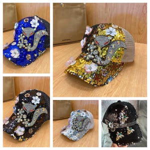 May include: A collection of sequined baseball caps in various colors, including blue, gold, black, and silver. Each cap features an embellished fox design with floral and crystal accents. The caps have a mesh back.