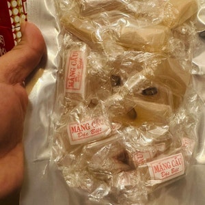 May include: A clear plastic bag filled with individually wrapped pieces of a light brown candy. The candy wrappers have red text that reads "MANG CẦU Đặc Biệt".