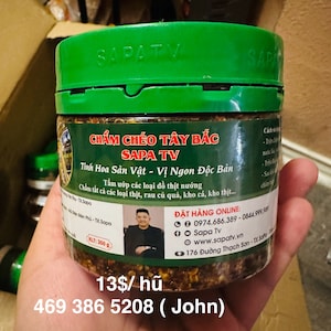 May include: A green jar with a white label that reads "Chẩm Chéo Tây Bắc Sapa TV" in Vietnamese. The label also includes the text "Tỉnh Hoa Sản Vật - Vị Ngon Độc Bản" in Vietnamese. The jar contains a brown and green mixture. The jar is sitting on a brown surface. The text "13$/hũ" is visible at the bottom of the image.