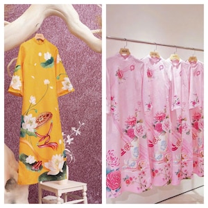 May include: A display of traditional Vietnamese áo dài dresses. One dress is a vibrant yellow with a floral design, while the others are pink with floral patterns. The dresses are hanging on a rack and a wooden branch.