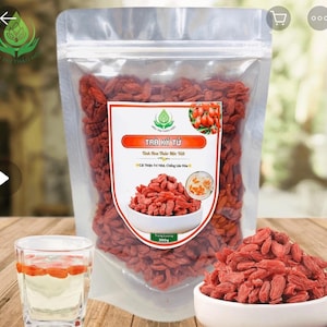 May include: A clear plastic bag filled with dried red goji berries, with a white label featuring text in Vietnamese. A small bowl of goji berries and a glass of goji berry tea are also visible. The bag contains 200g of berries.