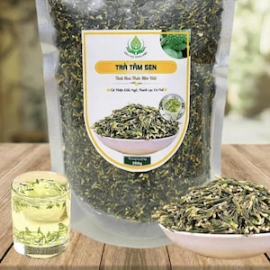 May include: A clear plastic pouch filled with dried green lotus plumule tea, labeled TRÀ TÂM SEN with a 200g weight. A small glass of brewed light yellow tea and a white bowl containing a pile of the dried green herbal tea leaves are in front.