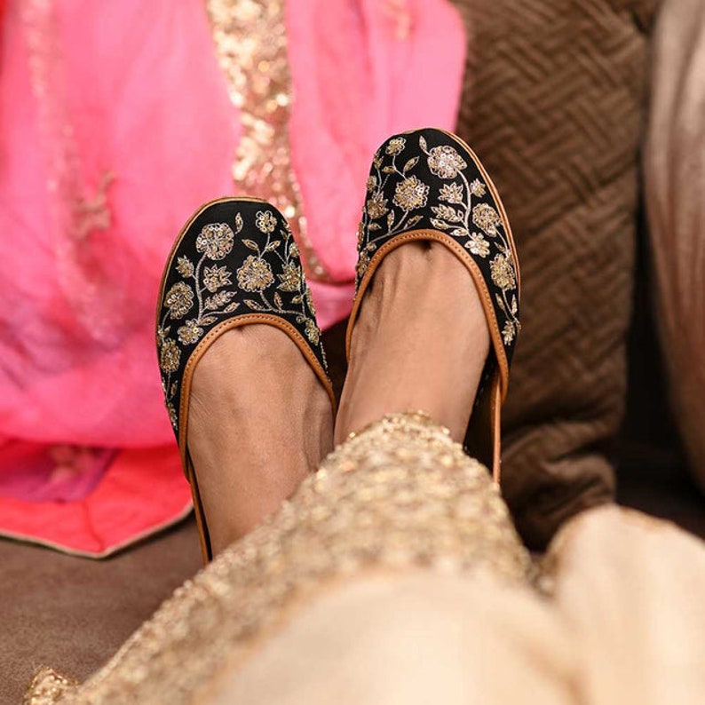 May include: A pair of black embroidered flat shoes with gold floral designs. The shoes are worn by a person wearing a gold and black outfit.