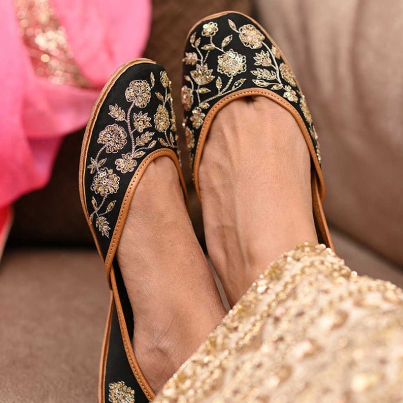 May include: A pair of black flat shoes with gold embroidery. The shoes have a pointed toe and a low heel. The embroidery features floral designs.