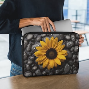 May include: A black laptop sleeve with a zipper closure features a sunflower with yellow petals and a dark brown center on a background of gray stones with water droplets.