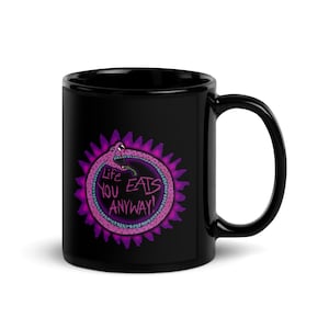 May include: A black ceramic mug featuring a graphic of a pink snake forming a circle while biting its own tail. Inside the circle, the text reads Life EATS YOU ANYWAY! in a stylized font. The design is surrounded by a purple spiky halo effect.