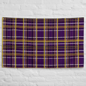 Puede incluir: Una bandera rectangular con un patrón de cuadros. El diseño presenta una cuadrícula de líneas que se cruzan en morado, marrón, blanco y amarillo. La bandera cuelga contra una pared de ladrillos blancos.