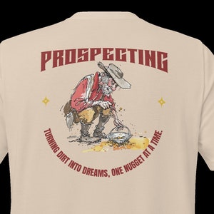 Peut inclure: Un t-shirt beige avec un graphique rouge d'un prospecteur de dessin animé en train de faire de l'or. Le graphique comprend le texte "Prospecting" et "Turning dirt into dreams, one nugget at a time."