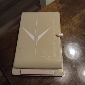 May include: Tan address book with a white geometric design and a sliding latch. The cover has the text "AREA CODES MEMO" and an alphabetical index along the side. The book is rectangular and appears to be made of metal.