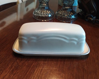 May include: A white ceramic butter dish with a decorative design and a light blue rim. The dish is sitting on a wooden table.