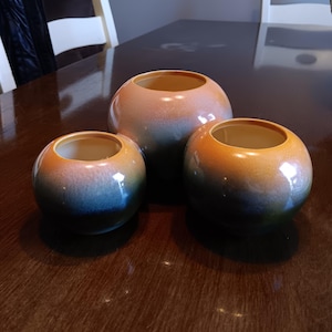 May include: Three ceramic pots with a gradient of orange, yellow, and blue. The pots are nested inside each other, with the largest pot at the bottom and the smallest pot at the top.