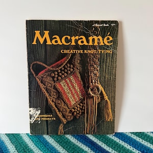 May include: A softcover instructional book titled Macramé Creative Knot-Tying with a dark wood background. The cover features a brown macramé bag with red and yellow accents and a long fringed strap with a gold metal ring, published by Sunset Books.