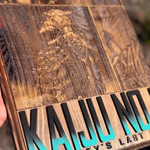 Puede incluir: Un letrero de madera con un dise&ntilde;o tallado y las palabras "KAIJU NO" en letras negras grandes con un degradado turquesa. Debajo, se muestra el texto "HUMANITY'S LAST". El letrero tiene un acabado marr&oacute;n oscuro.