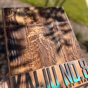 Puede incluir: Placa de madera con las palabras "KAIJU NO. 8" en letras negras y turquesas. La placa tiene un dise&ntilde;o tallado detallado y tambi&eacute;n son visibles las palabras "HUMANITY'S LAST STAR". La madera tiene un tono c&aacute;lido y natural.