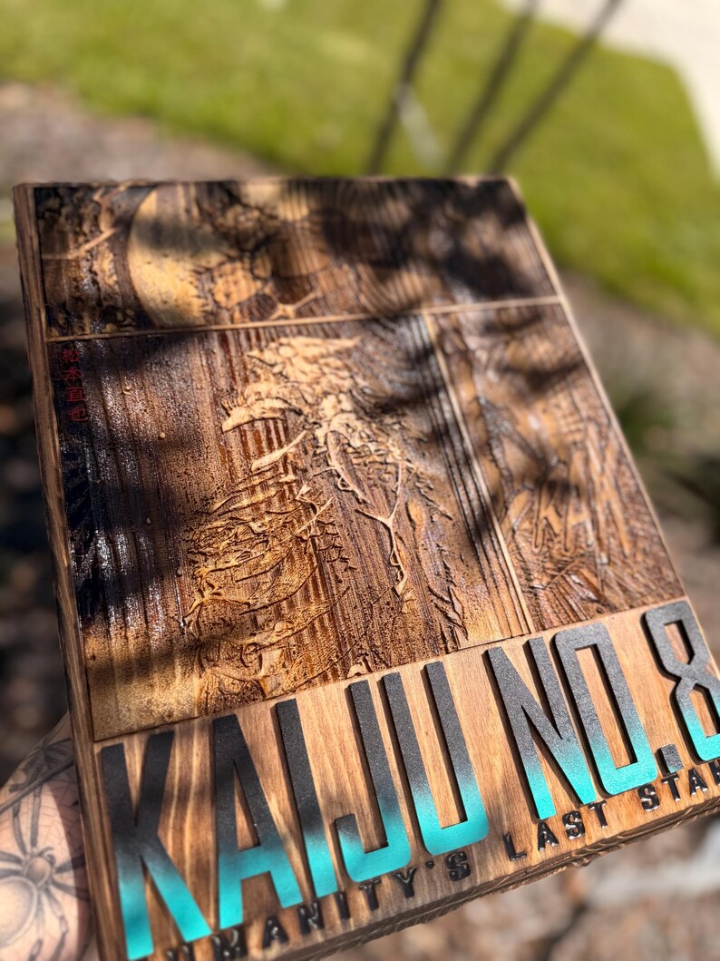 Puede incluir: Placa de madera con las palabras "KAIJU NO. 8" en letras negras y turquesas. La placa tiene un dise&ntilde;o tallado detallado y tambi&eacute;n son visibles las palabras "HUMANITY'S LAST STAR". La madera tiene un tono c&aacute;lido y natural.