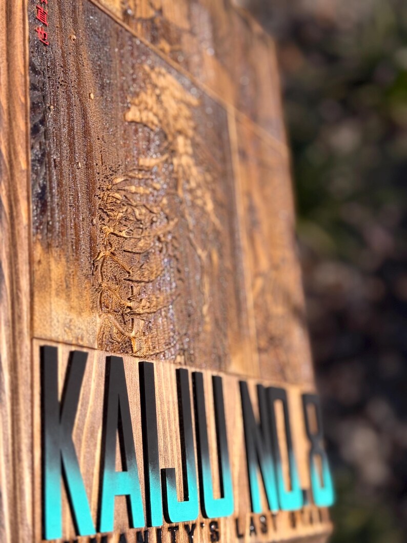 Puede incluir: Un letrero de madera con las palabras "KAIJU NO.8" en letras negras con un degradado turquesa. El letrero tiene un dise&ntilde;o tallado y el texto "HUMANITY'S LAST STAND" en la parte inferior. La madera tiene una veta natural y un tinte oscuro.
