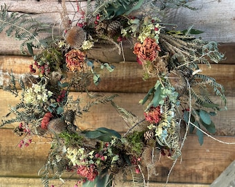 May include: A rustic wreath composed of dried flowers, berries, and greenery. The wreath features a variety of textures and colors, including shades of green, brown, red, and cream. It is displayed against a wooden background.