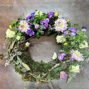 May include: A circular floral wreath featuring a variety of flowers in shades of purple, white, and green. The wreath includes dahlias, roses, and other blooms, with a base of moss, twigs, and small pinecones. The arrangement is set against a neutral background.