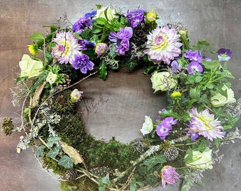 May include: A circular floral wreath featuring a variety of flowers in shades of purple, white, and green. The wreath includes dahlias, roses, and other blooms, with a base of moss, twigs, and small pinecones. The arrangement is set against a neutral background.