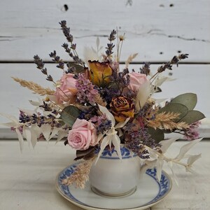 May include: Dried flower arrangement in a white teacup with a blue and white pattern. The arrangement includes pink roses, dried lavender, and other dried flowers.
