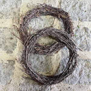 May include: Two brown twig wreaths, one smaller than the other, are stacked on top of each other on a light gray stone surface.