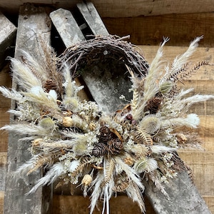 May include: A dried floral wreath with a natural twig base. The wreath is decorated with dried pampas grass, pine cones, and other dried botanicals in shades of beige, brown, and white. The wreath is hanging on a rustic wooden background.