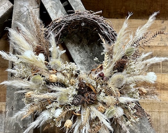 May include: A dried floral wreath with a natural twig base. The wreath is decorated with dried pampas grass, pine cones, and other dried botanicals in shades of beige, brown, and white. The wreath is hanging on a rustic wooden background.