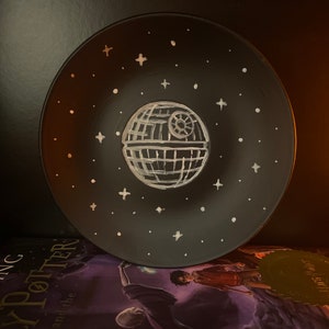 May include: A black ceramic plate with a white painted design of the Death Star from Star Wars. The plate is decorated with white stars.