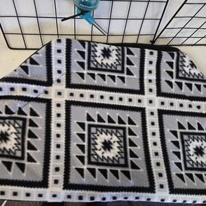 May include: A gray, black, and white patterned pet bed with a geometric design. The bed has a black border and is placed near a wire cage and a water bottle. The pattern includes squares, triangles, and star-like shapes.