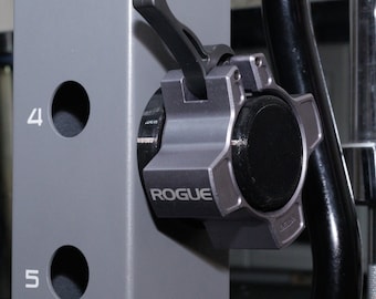 May include: A gray metal weight plate collar with a black lever and the word "ROGUE" printed on the side. The collar is attached to a gray metal weight rack.