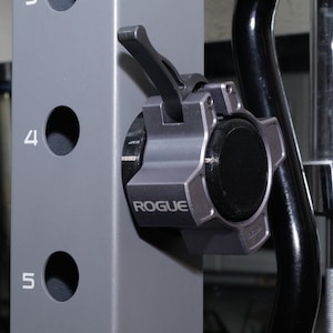 May include: A gray metal weight plate collar with a black lever and the word "ROGUE" printed on the side. The collar is attached to a gray metal weight rack.