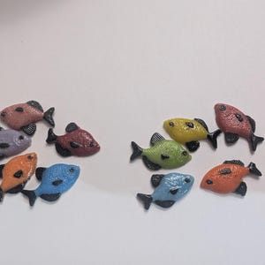 May include: A collection of colorful, miniature fish figurines. The fish are in various colors, including pink, purple, orange, blue, yellow, and green. Each fish has black fins and a textured surface, creating a playful and decorative aesthetic.