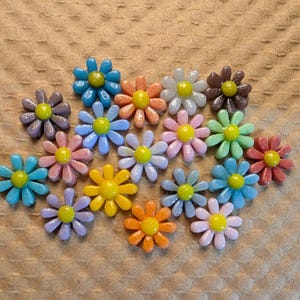 May include: A collection of colorful daisy-shaped embellishments in various shades of blue, pink, orange, yellow, green, and purple. Each flower has a yellow center. The embellishments are scattered on a textured, brown surface.