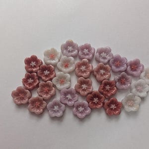 May include: A collection of small, flower-shaped beads in shades of pink, white, and purple. Each bead has a detailed petal design and a contrasting center, perfect for jewelry making or craft projects.