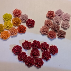 May include: A collection of small, three-dimensional rose-shaped cabochons arranged in three distinct clusters. The colors include various shades of yellow, orange, red, pink, and mauve, each featuring a glossy finish and detailed petal layers.