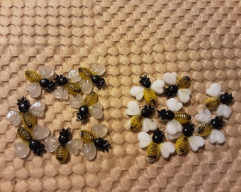 May include: Several small, decorative bee-shaped objects with yellow and black striped bodies, black heads, and white wings. The bees are arranged on a textured, honeycomb-patterned surface. The bees are likely used for crafting or decoration.