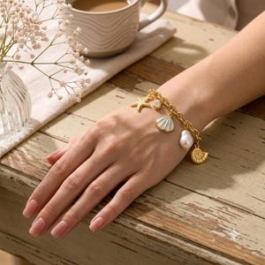 Peut inclure: Un bracelet à chaîne dorée orné de plusieurs breloques, dont une étoile de mer, une petite pierre transparente, un coquillage blanc, une perle blanche et un coquillage en spirale. Le bracelet est porté au poignet, révélant les maillons.