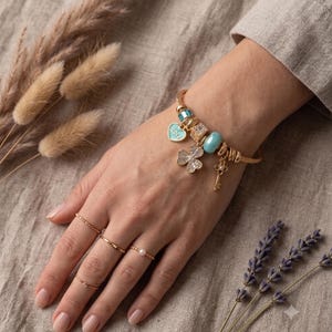 Peut inclure: Un bracelet à breloques doré orné d'un cœur bleu clair avec l'inscription Love, d'un trèfle scintillant, d'une petite clé et de perles bleues. Le bracelet est porté avec trois bagues dorées fines, dont l'une est agrémentée d'une petite perle blanche.