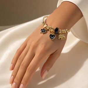 Peut inclure: Un bracelet à breloques doré composé d'une série de perles métalliques et de plusieurs pendentifs, dont un trèfle à quatre feuilles noir, un cœur en pierre noire, une perle noire à facettes et une figurine d'éléphant en or texturé.