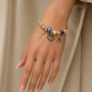 Peut inclure: Un bracelet à breloques doré orné de diverses perles et de pendentifs, notamment une clé, un coquillage, un cœur et une étoile de mer. Le bracelet comprend une perle bleue lisse et une perle bleu foncé, séparées par des intercalaires dorés.