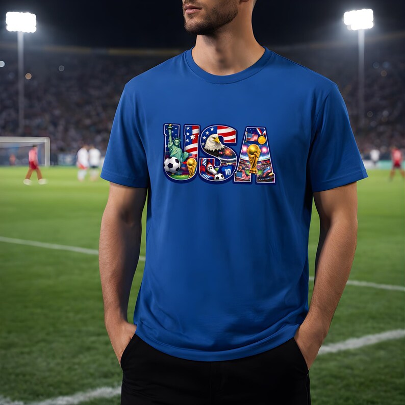 May include: Blue t-shirt with the word "USA" in a colorful design featuring the Statue of Liberty, an eagle, soccer balls, and the World Cup trophy. The shirt is short-sleeved.